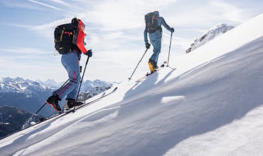 Zwei Personen fahren unter strahlend blauem Himmel einen schneebedeckten Berg hinauf. Sie sind in Winterkleidung mit Rucksäcken und Skistöcken gekleidet und haben einen Panoramablick auf die fernen schneebedeckten Gipfel.