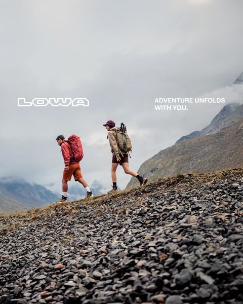 Zwei Wanderer gehen auf einem felsigen Hang in den Bergen unter einem bewölkten Himmel, mit dem Text "LOWA" und "ADVENTURE UNFOLDS WITH YOU.".